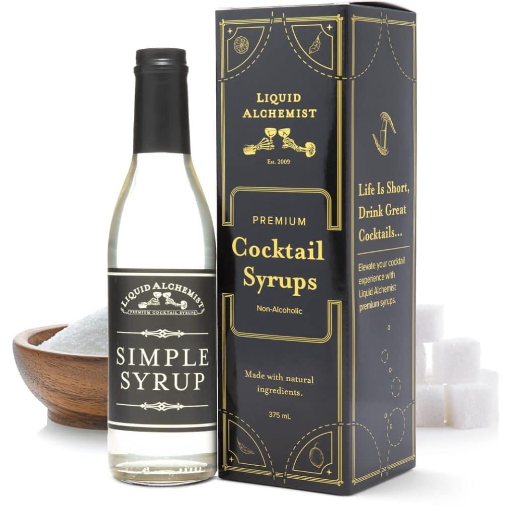 A bottle of Liquid Alchemist Simple Syrup sits elegantly before its packaging labeled "Premium Cocktail Syrups." Nearby, a wooden bowl and sugar cubes await their role in crafting delightful drinks. The label suggests, "Life is Short, Drink Great Cocktails," perhaps a strawberry basil cocktail awaits?.